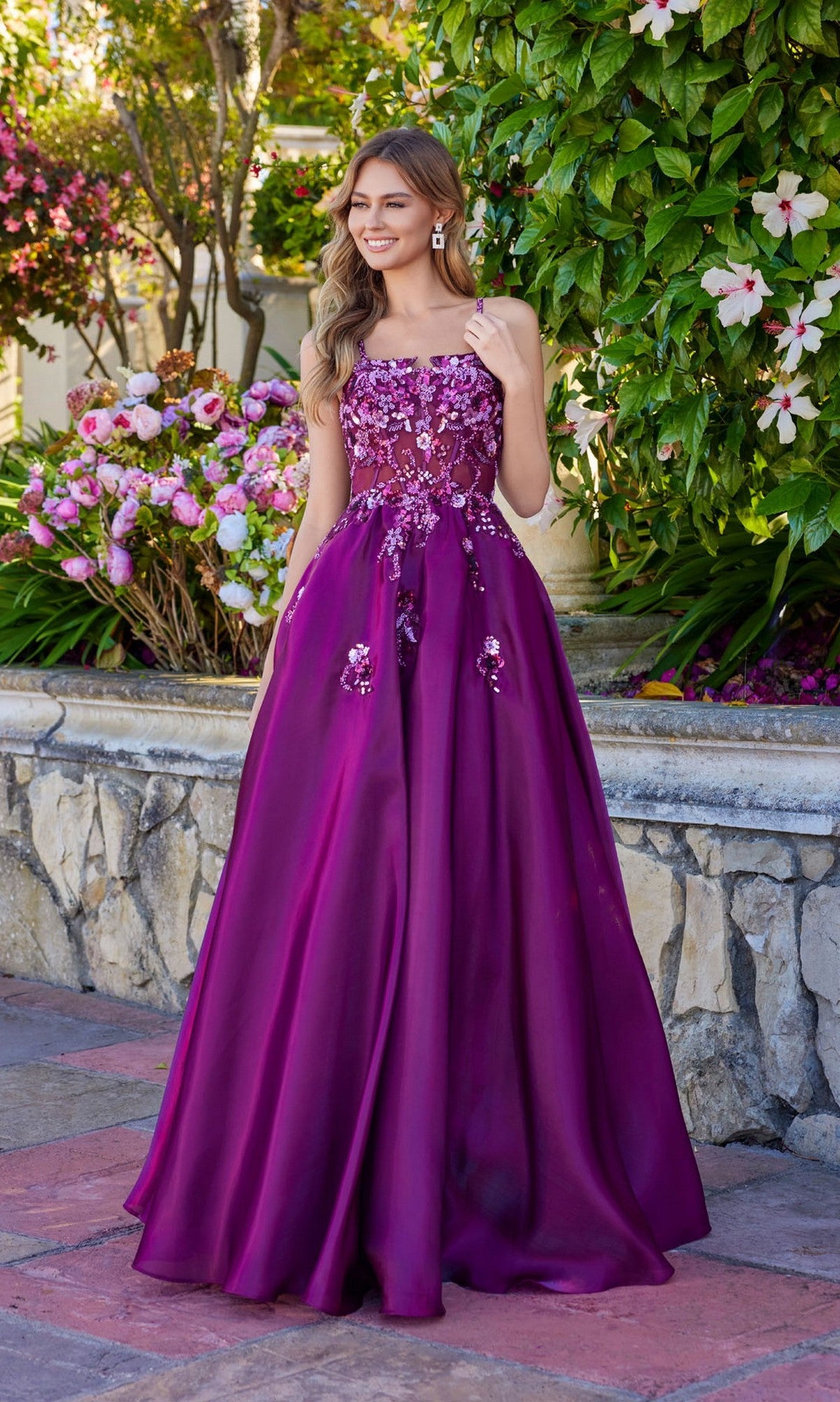 A woman stands outdoors in Juliet JT2617U, a strapless, floor-length magenta A-line prom dress with floral sequin embellishments and a lace-up back, surrounded by flowers and greenery.